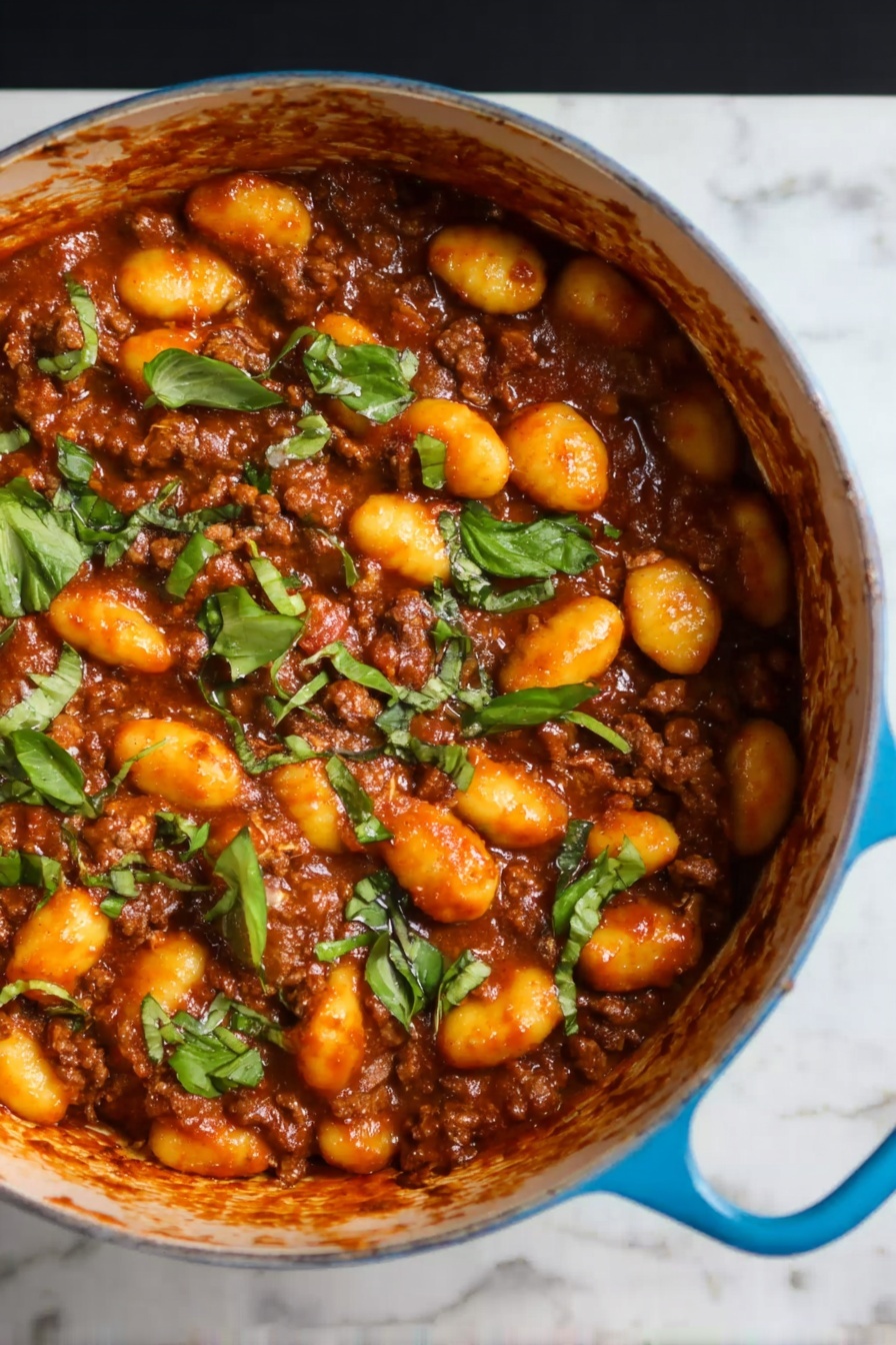 Gnocchi Bolognese, Italian gnocchi recipes, easy Bolognese sauce, comfort food dinner, hearty pasta dishes - The image shows a blue pot filled with a rich brown meat sauce mixed with soft, yellow-orange gnocchi. The top layer is scattered with pieces of fresh green herbs, adding a bright contrast to the warm colors of the sauce and gnocchi. The pot's handle is visible on the right side, resting on a white marbled surface. The texture of the gnocchi looks smooth and tender, while the meat sauce appears thick and hearty, covering all pieces evenly. Photo taken with an iphone --ar 2:3 --v 7