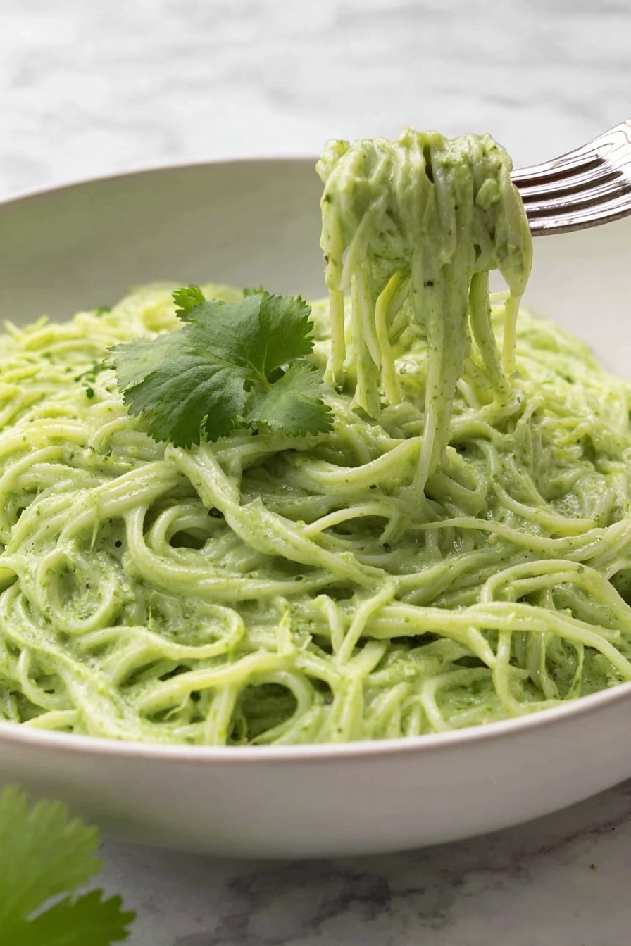 Vegan Cilantro Lime Noodles, vegan noodle recipes, cilantro lime pasta, easy vegan dinner ideas, quick healthy vegan meals - This image shows a white bowl filled with creamy green noodles, with about two layers of noodles visible, all coated evenly in a thick, smooth green sauce. On top of the noodles, there is a single thin slice of lime placed near the center, with bits of black pepper sprinkled across the noodles. Next to the lime slice, a small bunch of fresh green cilantro leaves rests gently. A silver fork is placed on the right edge of the bowl, partly inserted into the noodles. The bowl sits on a white marbled surface. Photo taken with an iphone --ar 2:3 --v 7