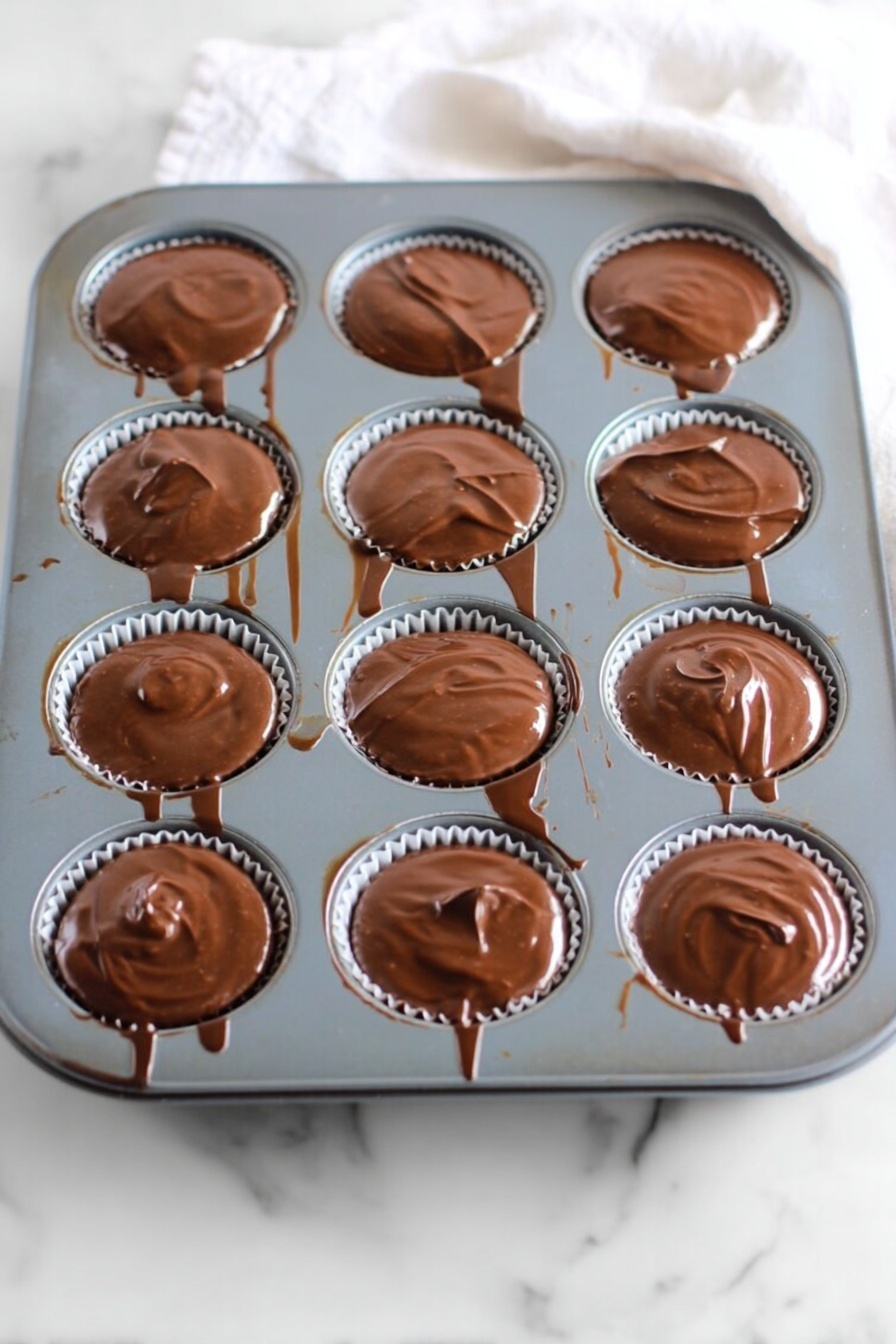 No Bake Peanut Butter Oat Cups, healthy peanut butter snack, easy no bake treats, quick oatmeal cups, chocolate peanut butter dessert - A gray muffin tray with twelve round cups, each filled with a smooth, glossy layer of melted chocolate that slightly spills over the edges. The chocolate has a shiny, swirled texture on top, and thin chocolate drips and streaks connect some of the cups and run down the tray’s sides. The tray rests on a white marbled surface with a soft white cloth partially visible at the top right corner. Photo taken with an iphone --ar 2:3 --v 7