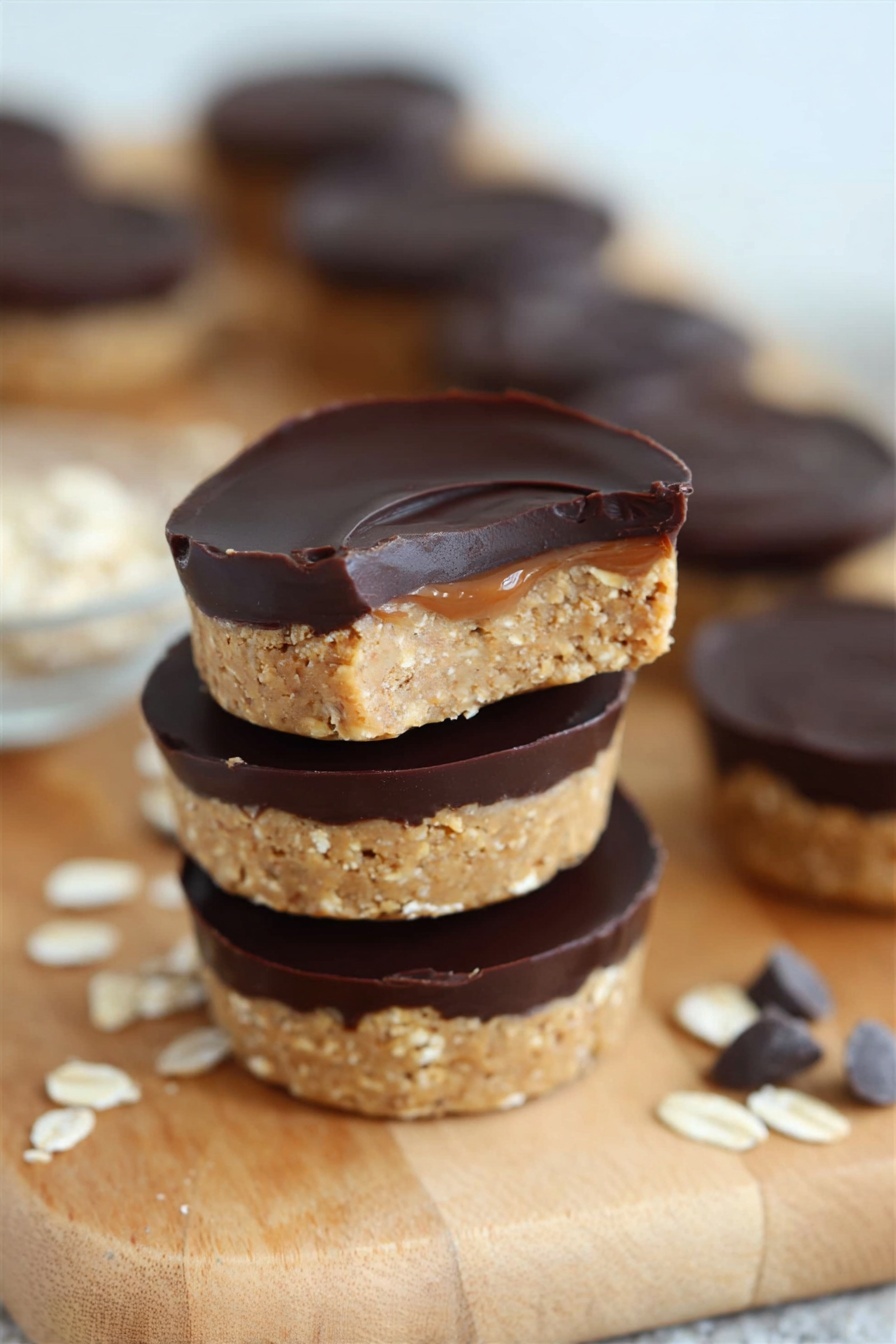 No Bake Peanut Butter Oat Cups, healthy peanut butter snack, easy no bake treats, quick oatmeal cups, chocolate peanut butter dessert - A close-up of a woman's hand holding a small round dessert with two layers, the bottom layer is a light tan, crumbly oat mixture, and the top layer is smooth, dark chocolate spread evenly. In the background, several more of these two-layer treats are stacked and scattered on a wooden board set on a white marbled surface, with some oats sprinkled nearby. The overall scene is clean and bright with soft lighting, highlighting the texture contrast between the oat base and the chocolate top. Photo taken with an iphone --ar 2:3 --v 7