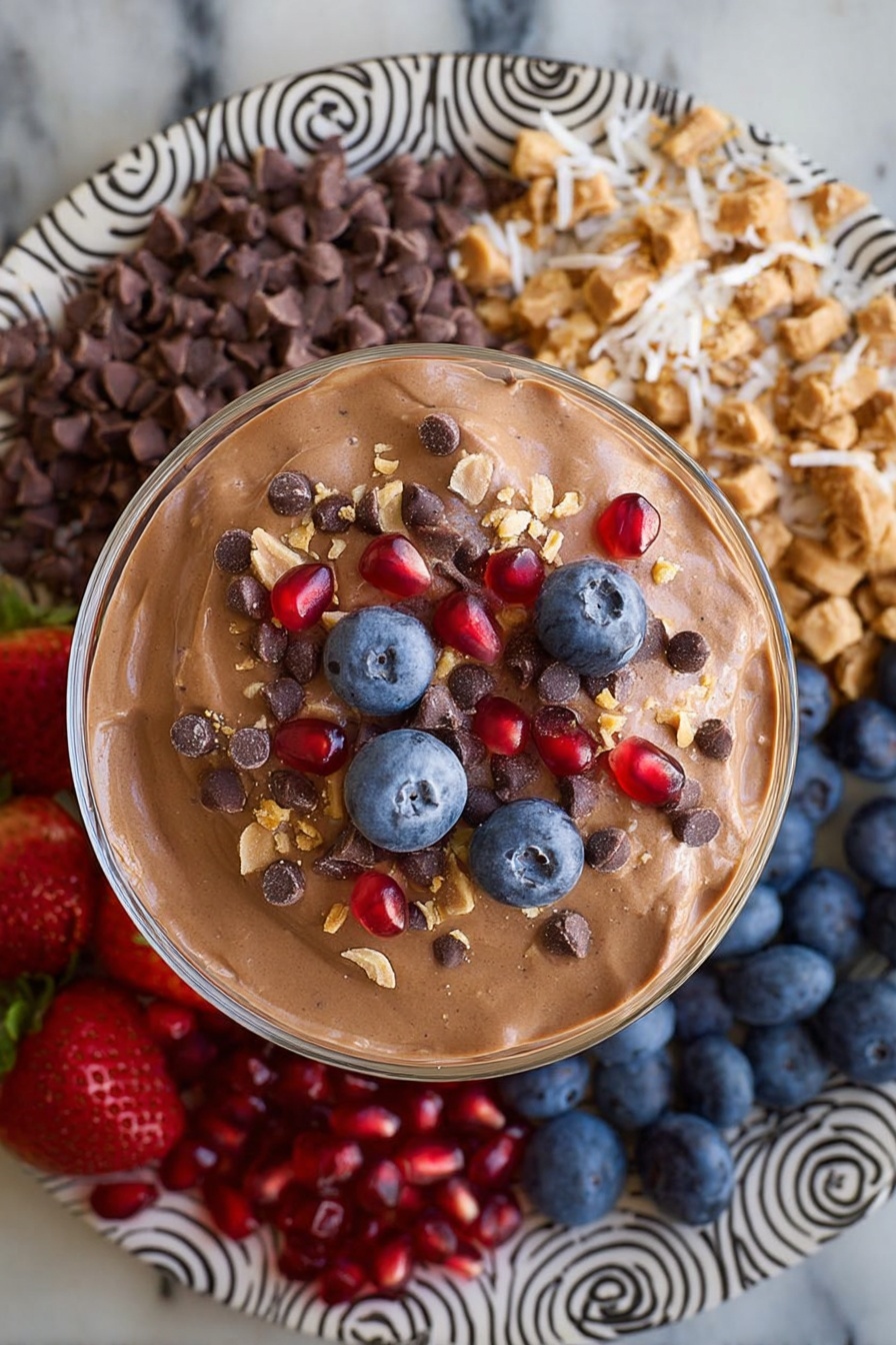 Chocolate Protein Fluff, healthy chocolate protein dessert, high-protein fluffy snack, quick protein dessert, low-sugar chocolate treat - A large round white plate with black spiral and zigzag patterns holds eight sections of colorful food arranged around a central glass bowl filled with smooth light brown chocolate mousse. Starting from the top left going clockwise, there are bright red whole strawberries with green tops, small dark brown chocolate chips, off-white shredded coconut, chopped pieces of chocolate peanut butter cups, bright red pomegranate seeds, dark blue blueberries, beige crunchy granola clusters, and then back to the strawberries. The plate is placed on a white marbled surface. Photo taken with an iphone --ar 2:3 --v 7