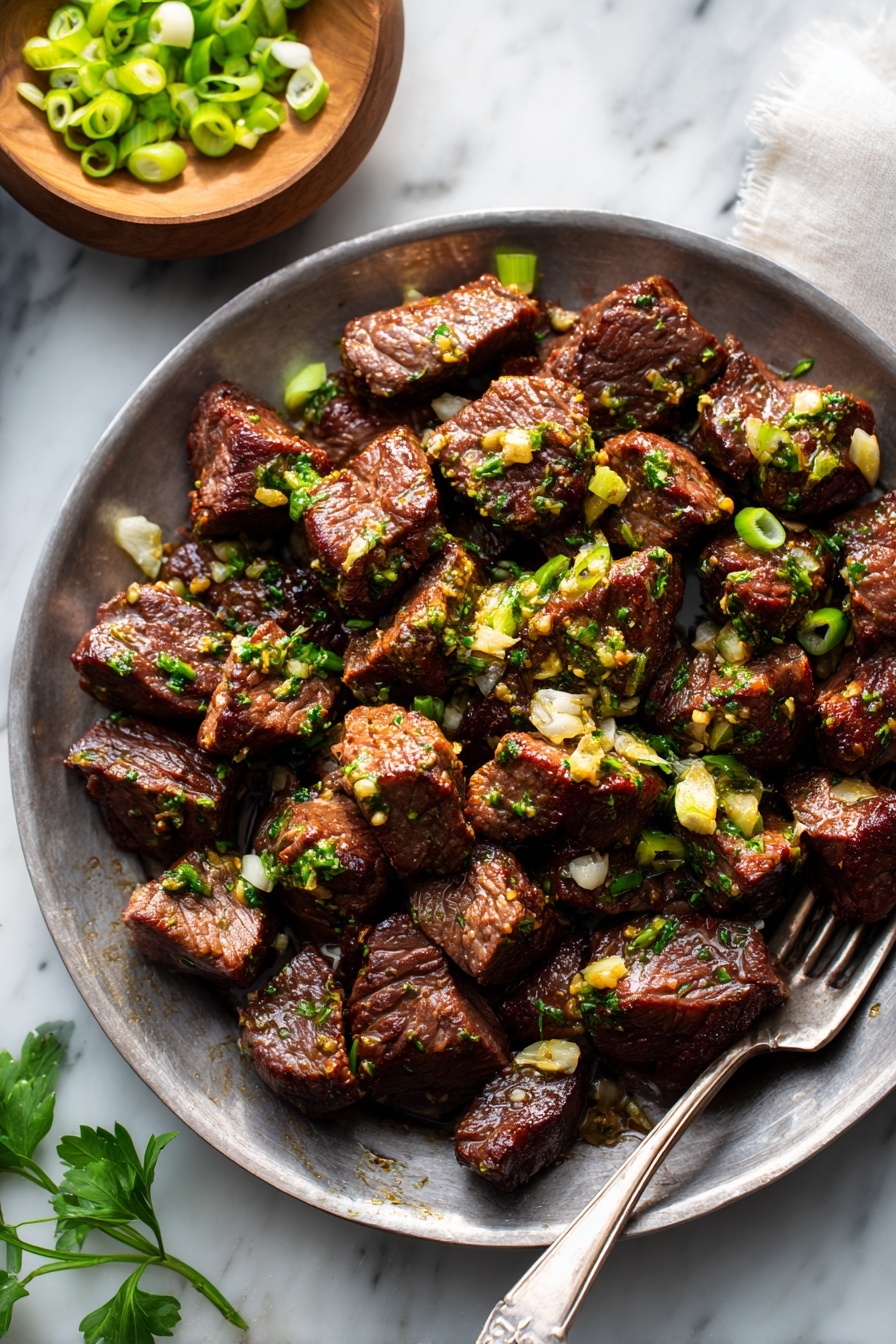 Garlic Butter Steak Bites, steak bite recipes, quick steak dinner, easy steak recipes, tender steak tips - A round plate full of small, browned meat pieces seasoned with chopped green herbs and garlic bits, scattered with thinly sliced light green onion rings on top. The plate is resting on a white marbled surface with some parsley leaves and a fork nearby, and a small white bowl with more green onions is partly visible above the plate. The meat pieces have a textured, slightly glossy look from the seasoning. photo taken with an iphone --ar 2:3 --v 7