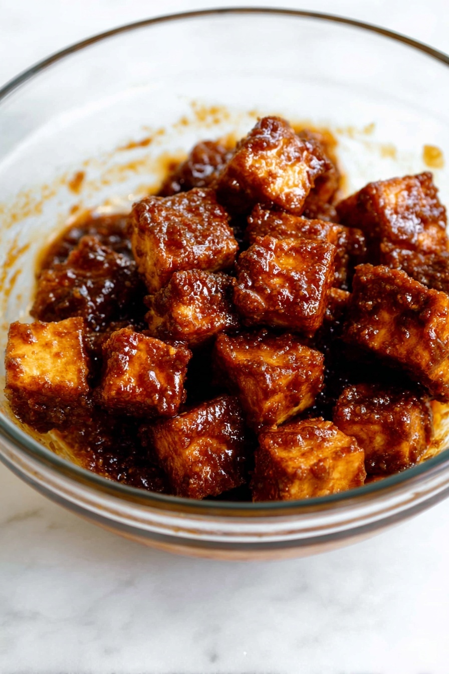 Crispy Gochujang Korean Tofu, Korean tofu recipe, spicy crispy tofu, gochujang tofu, healthy tofu dinner - The image shows a clear glass bowl filled with several small cubes of fried tofu. Each tofu piece is coated in a dark brown, shiny sauce with a textured surface that looks sticky and flavorful. The tofu pieces are piled together in the bowl, some slightly stacked on top of others. The smooth surface of the glass bowl lets you see the sauce clinging to the sides. The bowl is placed on a white marbled surface. photo taken with an iphone --ar 2:3 --v 7