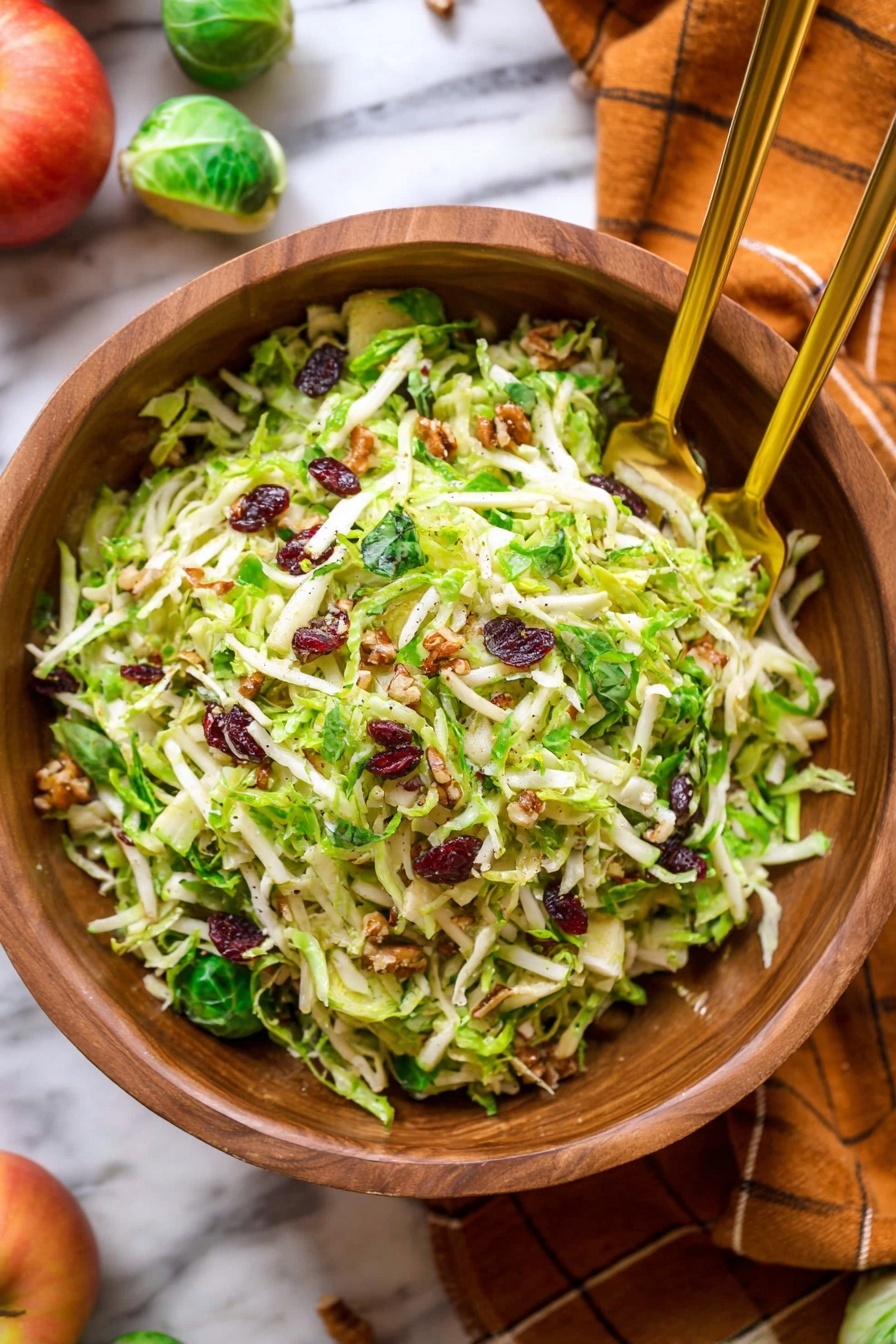 Kale Brussels Sprout Apple Salad with Bacon, healthy kale apple salad, savory Brussels sprout salad, easy bacon salad recipe, crunchy fruit and veggie salad - The image shows a wooden bowl full of a shredded green salad mixed with small bits of white cheese and dark red pieces, likely dried cranberries or bacon bits, with a texture of thin green leaves and stems. Two gold spoons rest in the bowl, positioned diagonally and partially buried in the salad. The bowl sits on a beige cloth over a white marbled surface, and in the background, bright orange small pumpkins and a few green Brussels sprouts are scattered on the same white marbled surface. The scene has a warm, natural feel with soft lighting. photo taken with an iphone --ar 2:3 --v 7
