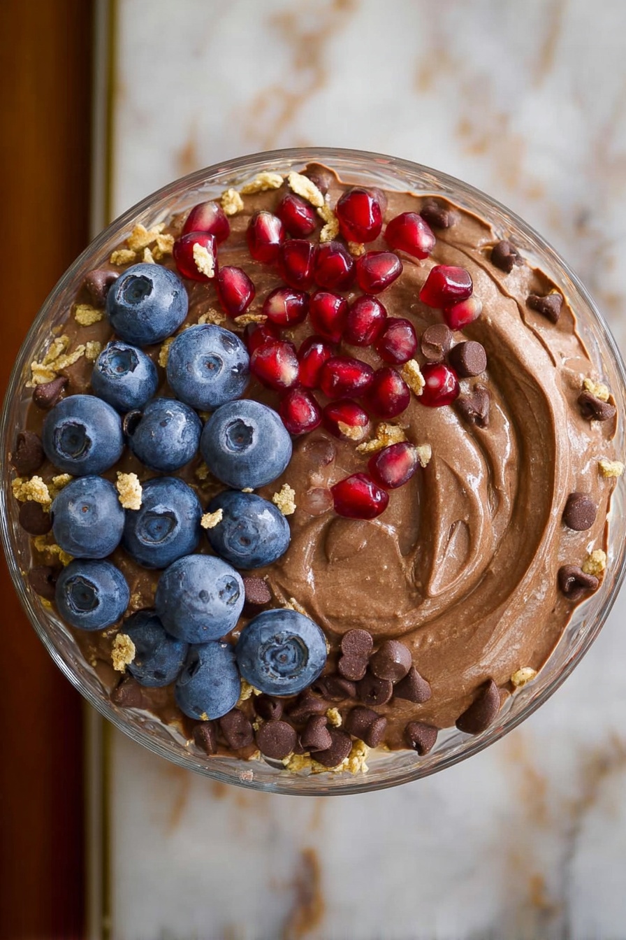 Chocolate Protein Fluff, healthy chocolate protein dessert, high-protein fluffy snack, quick protein dessert, low-sugar chocolate treat - A clear glass bowl in the center holds a creamy, smooth light brown pudding topped with a few scattered dark chocolate chips, bright red pomegranate seeds, fresh blue blueberries, and small crumbles of light tan granola. Surrounding the bowl on a white plate with black spiral and heart patterns are evenly placed piles of dark brown chocolate chips, white shredded coconut, light brown chopped peanut butter candy pieces, bright red strawberries with green tops, a heap of beige granola clusters, dark blue blueberries, and shiny red pomegranate seeds. The scene is set on a white marbled surface. photo taken with an iphone --ar 2:3 --v 7
