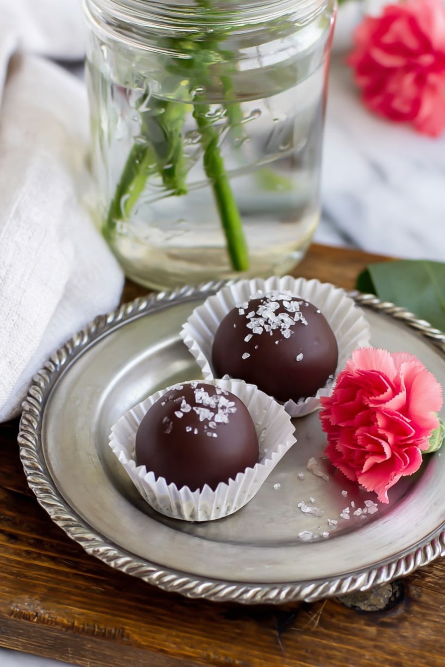 Strawberry Buttercream Candy Bites, strawberry candy treats, homemade strawberry confections, easy berry candies, no-bake strawberry sweets - The image shows two round chocolate balls on a small silver plate with scalloped edges. Each chocolate ball is smooth and dark brown, topped with white sugar crystals. One chocolate ball sits in a white paper cup liner, while the other lies directly on the plate. Next to the chocolates is a small bright pink carnation flower with a green stem, slightly touching the plate. The plate is placed on a wooden surface with a white marbled texture background partly visible. A clear glass jar with water and green stems is positioned behind the plate, and part of a white cloth napkin is seen near the jar photo taken with an iphone --ar 2:3 --v 7