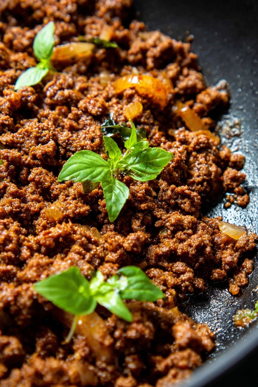 Thai Basil Beef Wraps, Thai Basil Beef Wraps recipe, easy Thai beef wraps, quick Thai beef meal, flavorful Thai beef wraps - This close-up image shows a single layer of cooked ground meat, rich brown in color with a slightly crumbly texture, mixed with small pieces of cooked onions that are golden yellow and translucent. Two small green basil sprigs with fresh leaves sit on top of the meat, adding a pop of bright green color. The food is in a black pan, which contrasts with the bright colors of the ingredients. The cooking oil shines slightly on the pan's surface, making the beef look moist and flavorful. Photo taken with an iphone --ar 2:3 --v 7