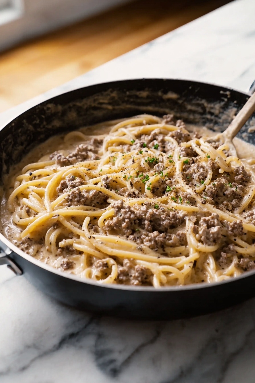 Creamy Beef and Garlic Butter Pasta, easy beef pasta recipe, one-pot creamy pasta, garlic butter pasta dinner, hearty weeknight pasta - A large metal pan sits on a gas stove with a sauce made of light brown creamy liquid, small clumps of browned ground meat, and short pale yellow pasta pieces scattered throughout. In the center, there is a small pile of finely grated pale yellow cheese. A wooden spoon rests on the edge of the pan, extending toward a wooden cutting board on the side. The background shows white ceramic jars on a white marbled surface beneath a window. Photo taken with an iphone --ar 2:3 --v 7