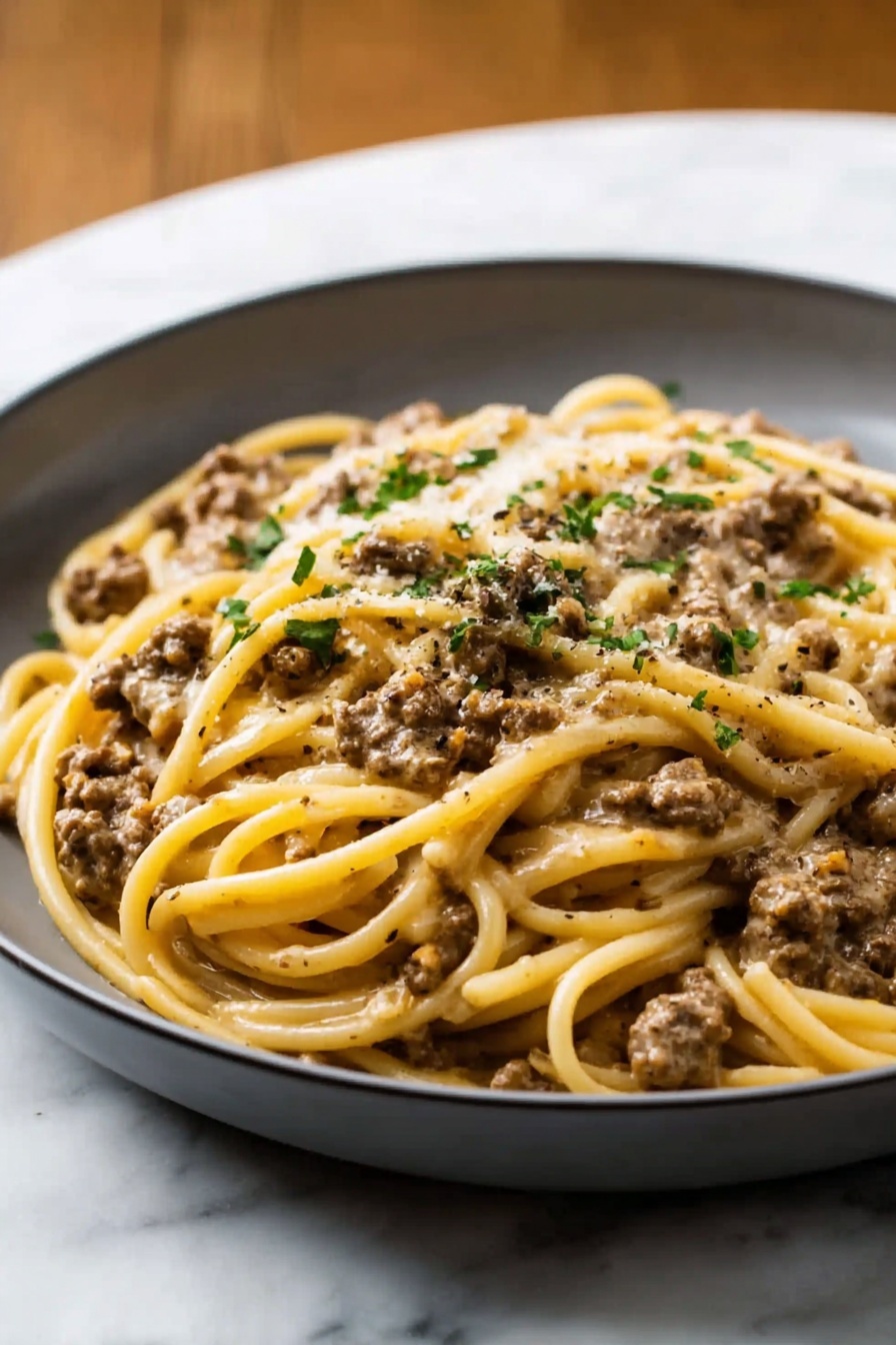 Creamy Beef and Garlic Butter Pasta, easy beef pasta recipe, one-pot creamy pasta, garlic butter pasta dinner, hearty weeknight pasta - A white shallow bowl filled with creamy pasta, showing one main layer of yellowish fettuccine noodles mixed with small light brown pieces of ground meat evenly spread throughout. The pasta is covered in a light beige creamy sauce, topped with a sprinkling of green chopped herbs and melted white cheese. The bowl sits on a wooden table next to a beige cloth with a fork beside it, and the background features a soft white marbled texture. photo taken with an iphone --ar 2:3 --v 7